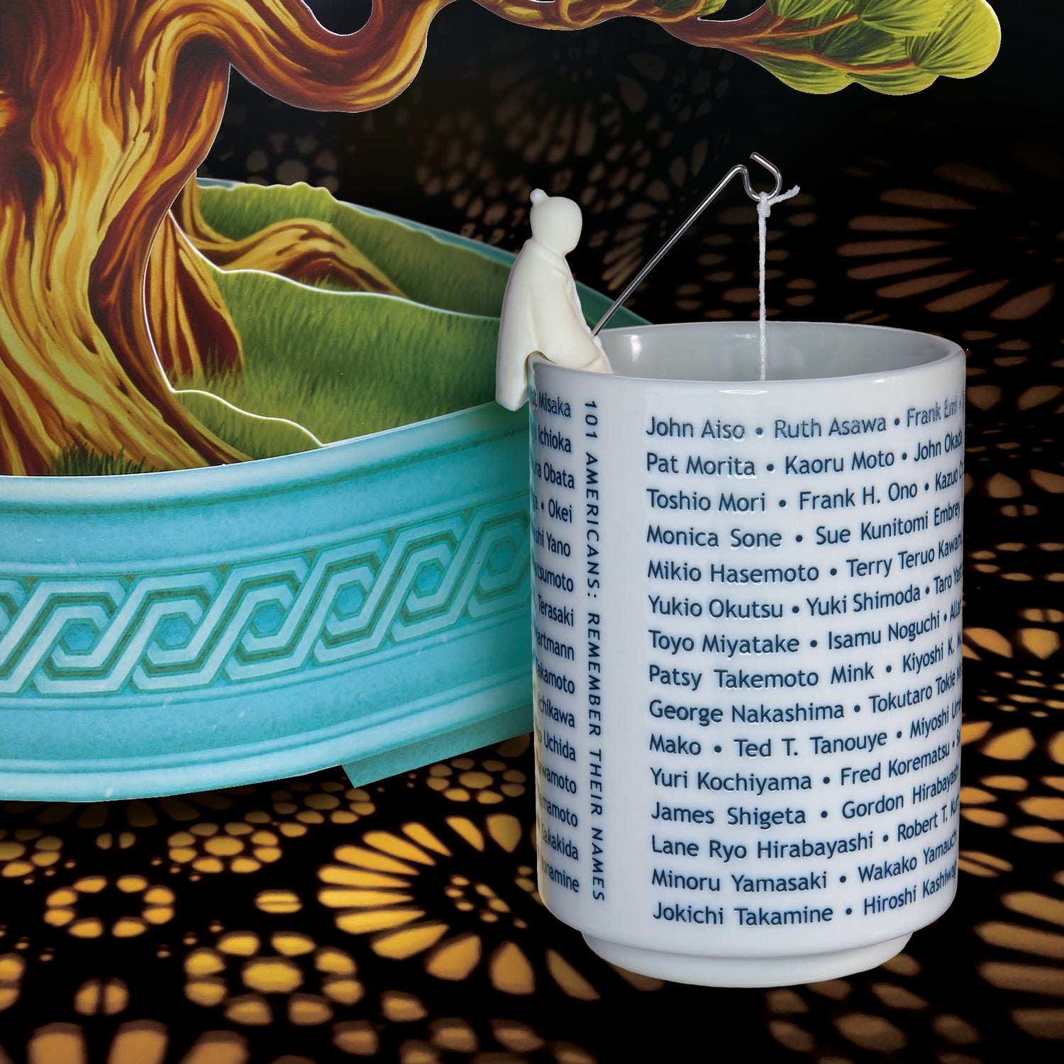 Large tea cup engraved with names of famous Japanese Americans. A small fisherman sits on the edge of the cup with a pole and a tea bag hanging from it. Close up pf a bonsai tree pop-up card in the background.