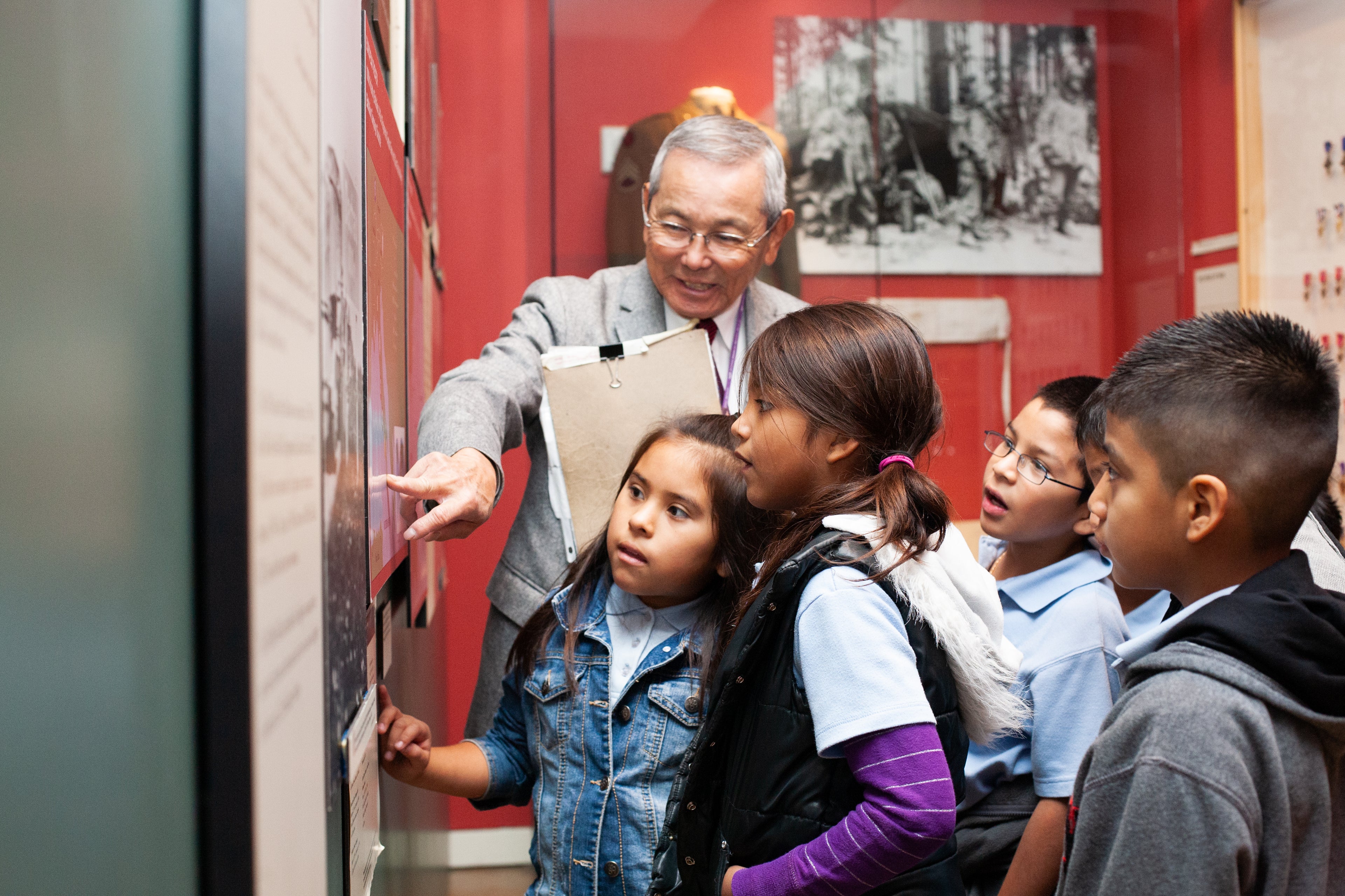 A group of children and an older man are gathered around a display board in the Japanese American National Museum with the man pointing at a panel while the children listen attentively.